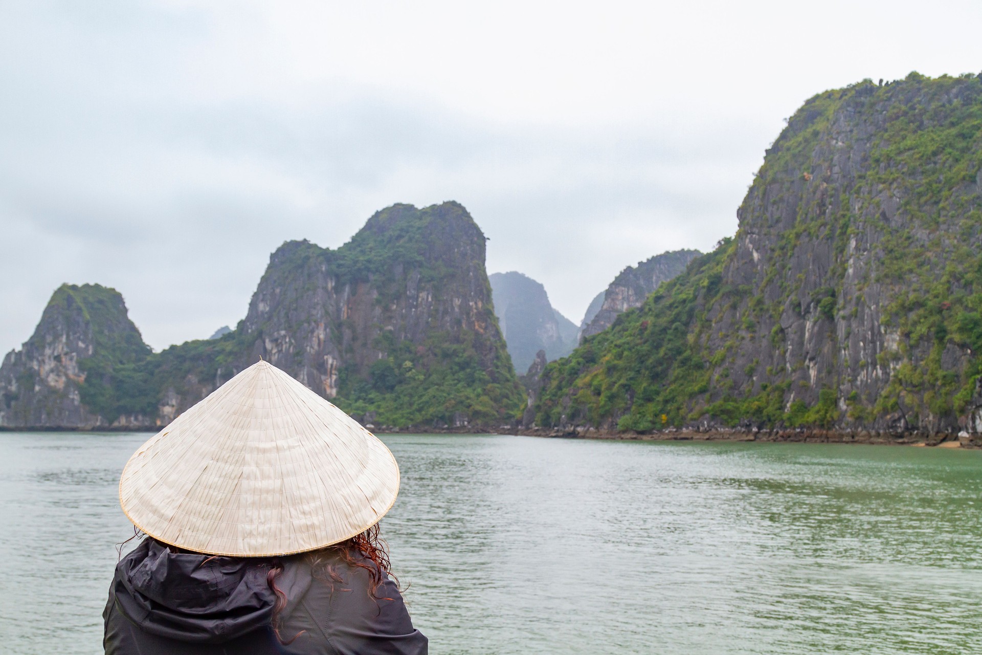 Mid adult female backpacker latin tourist wearing a traditional vietnamese non la conical hat is admiring the beautiful landscape of Ha Long Bay, an Unesco world heritage site and popular travel destination in Vietnam, during a boat trip in a cloudy raini