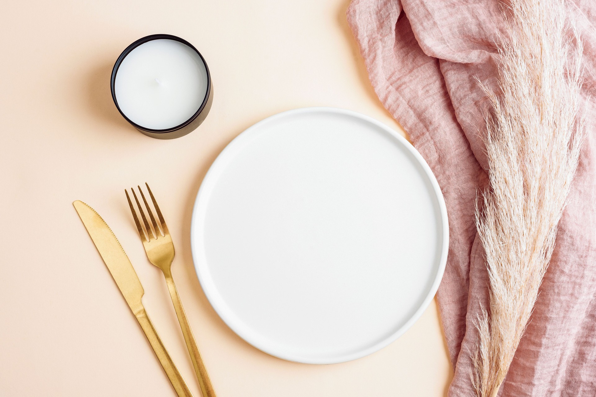 Flat lay of an elegant table setting with a white plate, golden cutlery, a pink linen napkin, and dried pampas grass on a soft beige background. Top view, mockup